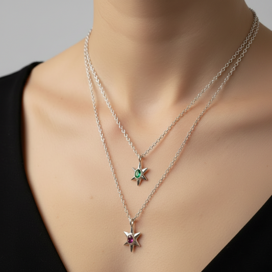 A close-up of a model's neck and chest wearing two layered silver chain necklaces with star pendants. The shorter necklace has a blue sapphire gemstone, and the longer necklace has a pink sapphire gemstone.
