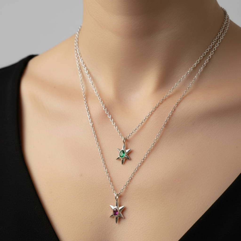 A close-up of a model's neck and chest wearing two layered silver chain necklaces with star pendants. The shorter necklace has a blue sapphire gemstone, and the longer necklace has a pink sapphire gemstone.