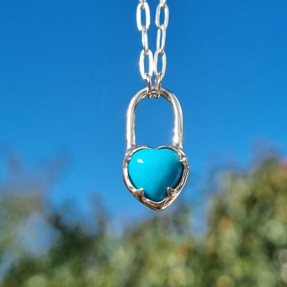 Turquoise cabochon heart pendant on a rectangle silver chain against a blue sky with greenery