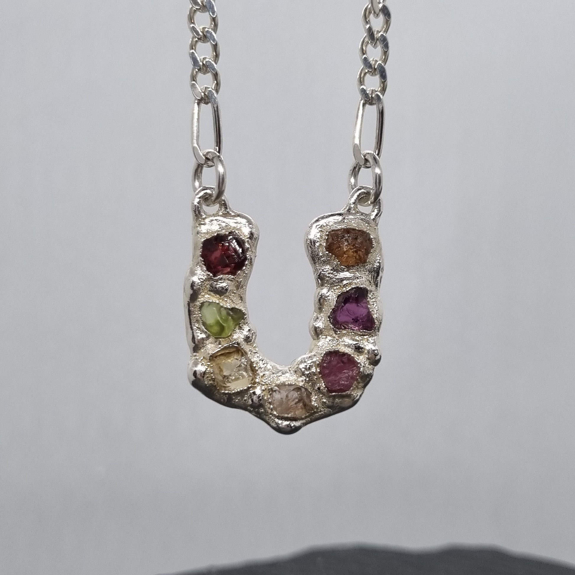 Sterling silver horseshoe pendant with embedded colourful raw natural gemstones on a figaro chain against a grey background. Handmade by Jo Cooper Jewellery