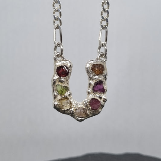Sterling silver horseshoe pendant with embedded colourful raw natural gemstones on a figaro chain against a grey background. Handmade by Jo Cooper Jewellery