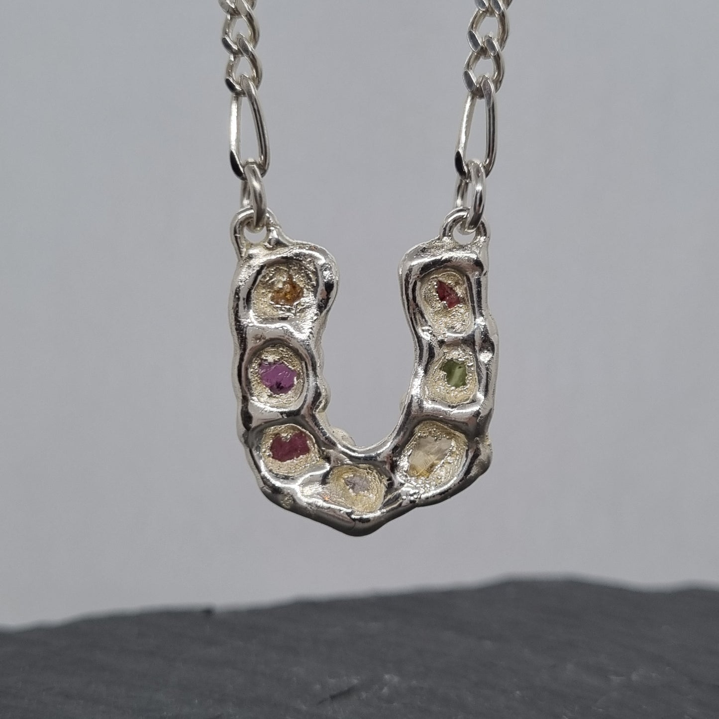 Back of sterling silver horseshoe pendant with embedded colourful raw natural gemstones on a figaro chain against a grey background