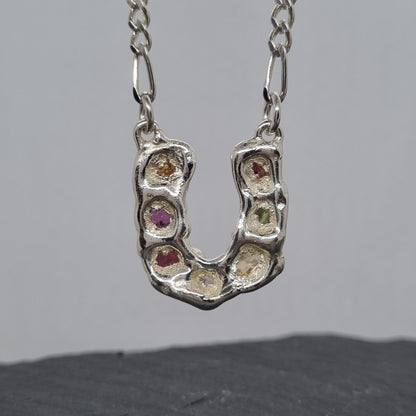 Back of sterling silver horseshoe pendant with embedded colourful raw natural gemstones on a figaro chain against a grey background