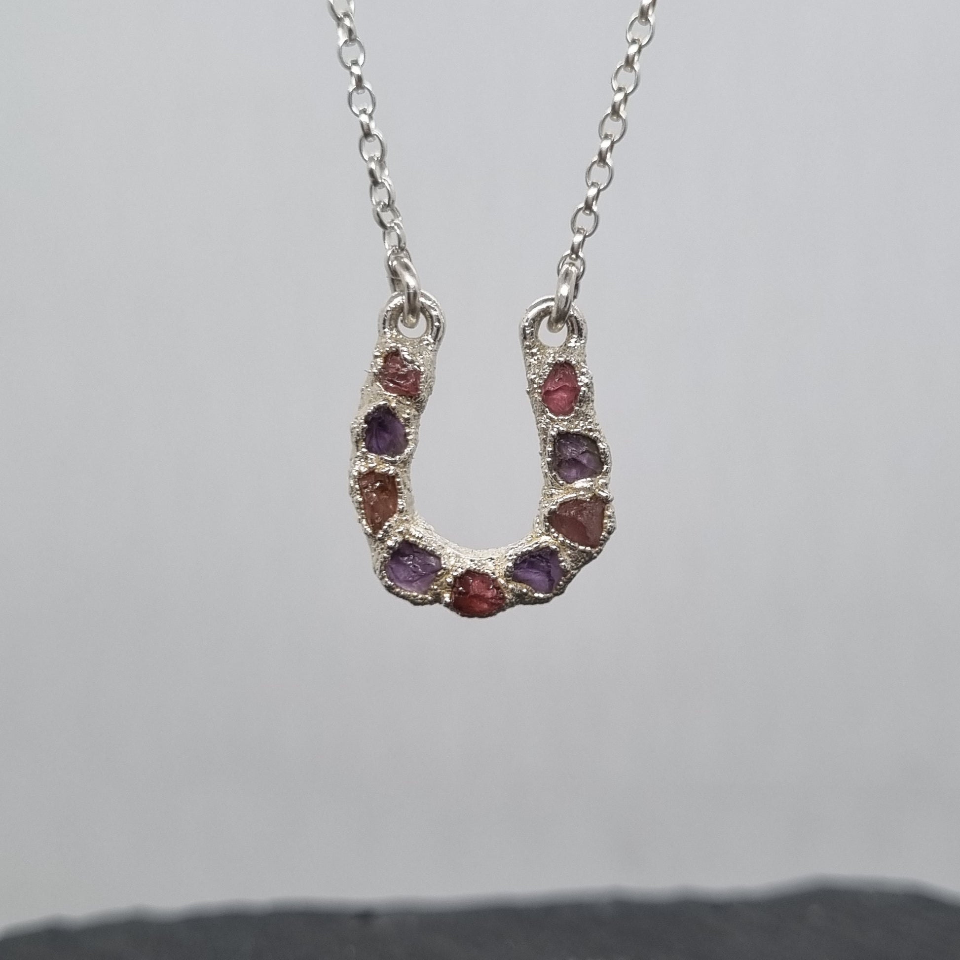 Sterling silver lucky horseshoe necklace with a pendant featuring raw pink tourmaline and purple amethyst gemstones set into a textured surface. Lunar New Year of the Horse Collection