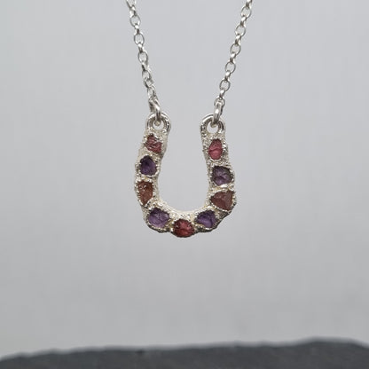 Sterling silver lucky horseshoe necklace with a pendant featuring raw pink tourmaline and purple amethyst gemstones set into a textured surface. Lunar New Year of the Horse Collection