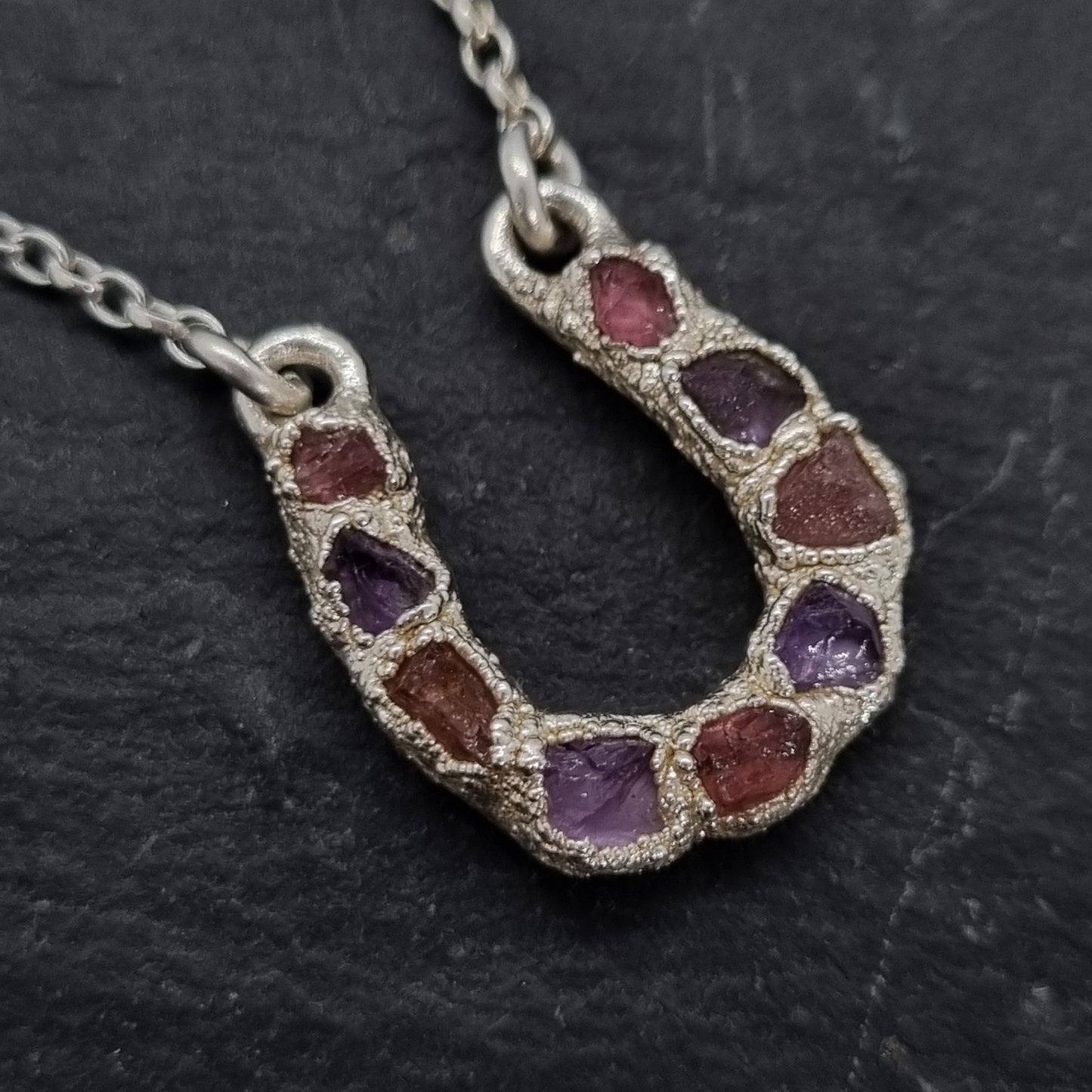 Close up of sterling silver lucky horseshoe necklace with a pendant featuring rough pink tourmaline and purple amethyst natural gemstones set into an electroformed textured surface. 