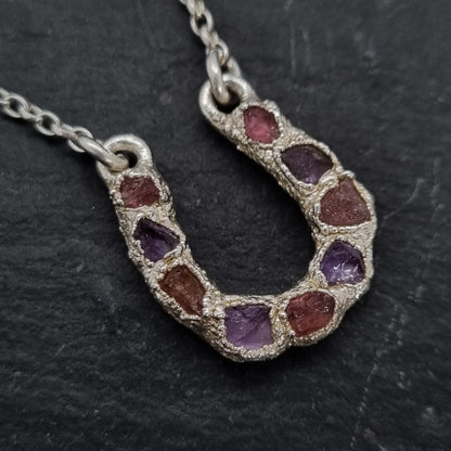 Close up of sterling silver lucky horseshoe necklace with a pendant featuring rough pink tourmaline and purple amethyst natural gemstones set into an electroformed textured surface. 