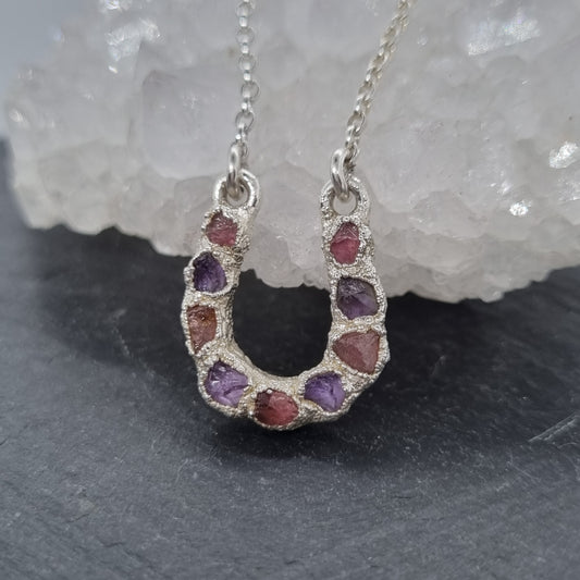 Sterling silver lucky horseshoe necklace with a pendant featuring raw pink tourmaline and purple amethyst gemstones set into a textured surface. Handmade by Jo Cooper Jewellery