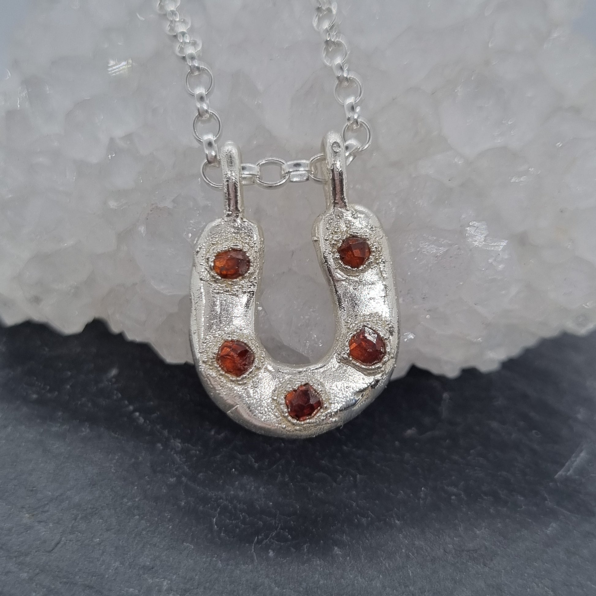 Handmade sterling silver horseshoe necklace with five raw garnet gemstones set in it. The silver has a rustic texture to it. On a silver belcher chain