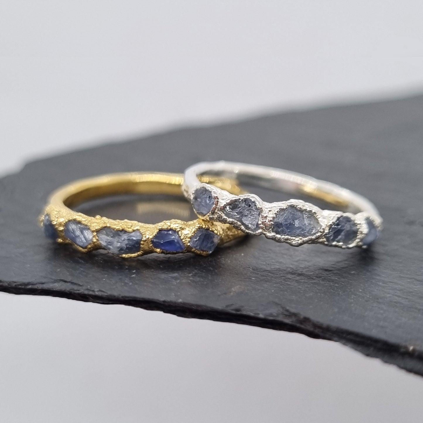Two rings, one silver one gold, with raw blue sapphire gemstones. The rings are textured. Displayed on a dark surface