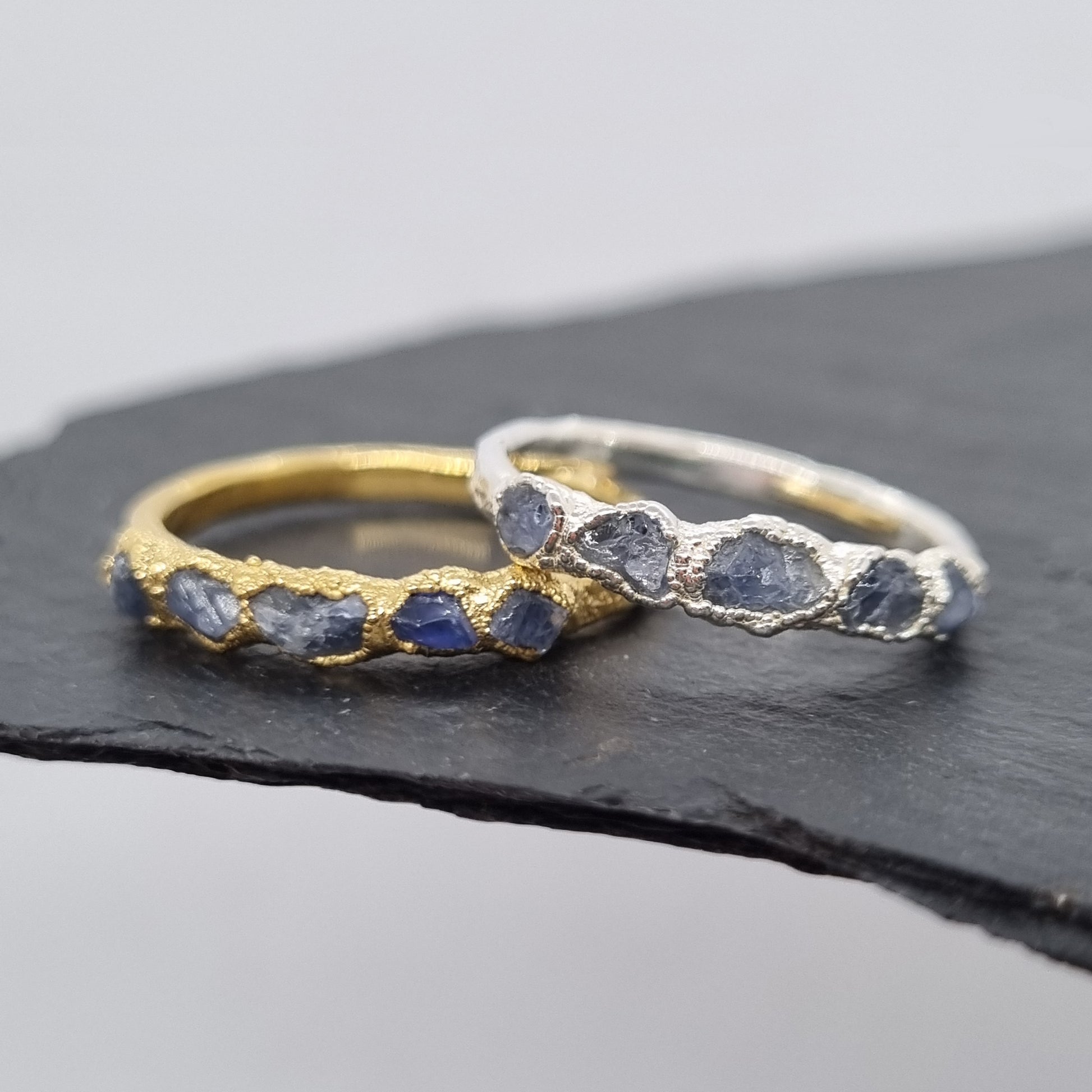 Two rings, one silver one gold, with raw blue sapphire gemstones. The rings are textured. Displayed on a dark surface