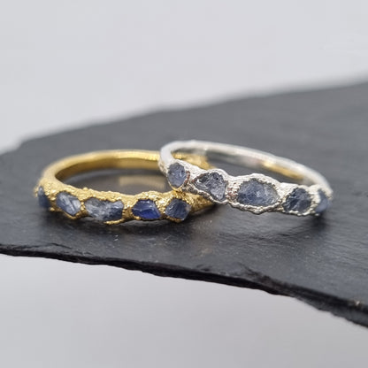 Two rings, one silver one gold, with raw blue sapphire gemstones. The rings are textured. Displayed on a dark surface