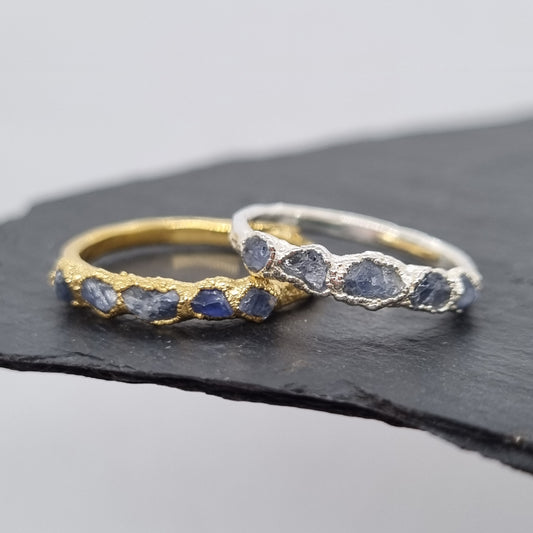 Two rings, one silver one gold, with raw blue sapphire gemstones. The rings are textured. Displayed on a dark surface