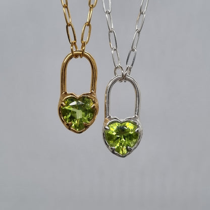 Two heart-shaped green peridot gemstone pendants on silver and gold chains against a gray background.
