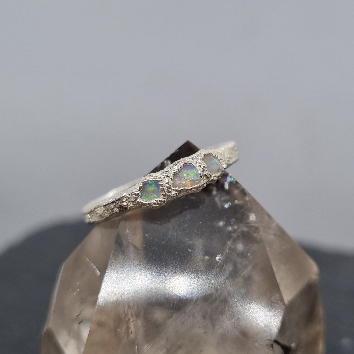 A fine silver ring with three freeform opal gemstones, shown close-up against a soft grey background with a translucent crystal prop.