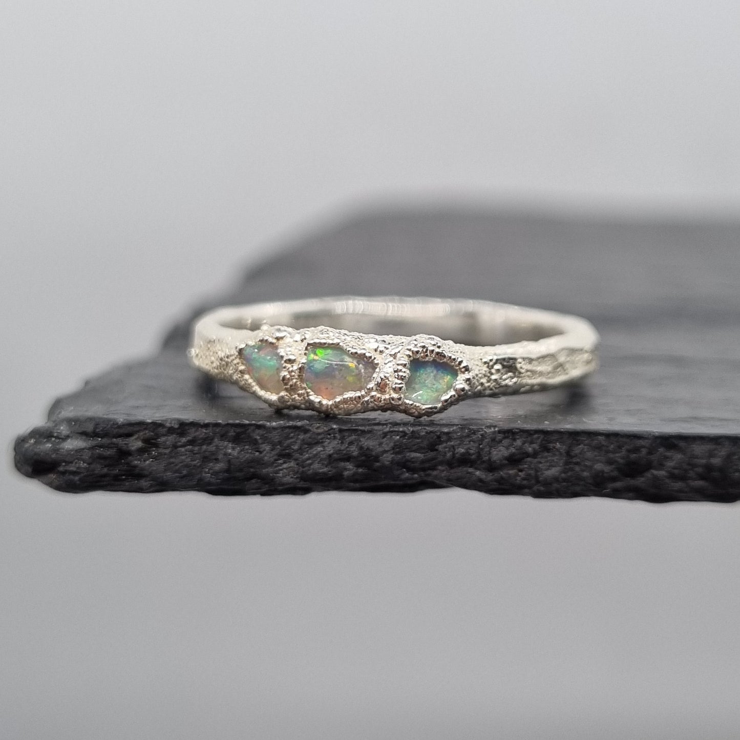 A silver ring with three small iridescent opal gemstones, displayed on a dark textured slate stand against a light gray background.