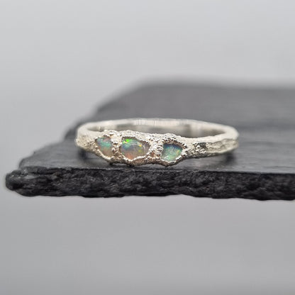 A silver ring with three small iridescent opal gemstones, displayed on a dark textured slate stand against a light gray background.