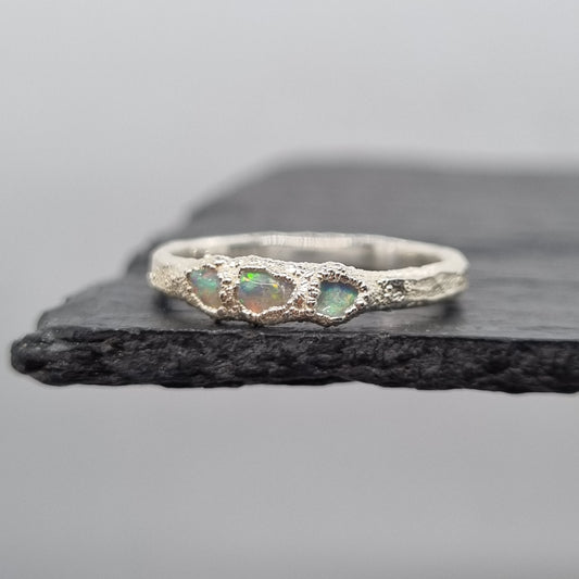A silver ring with three small iridescent opal gemstones, displayed on a dark textured slate stand against a light gray background.