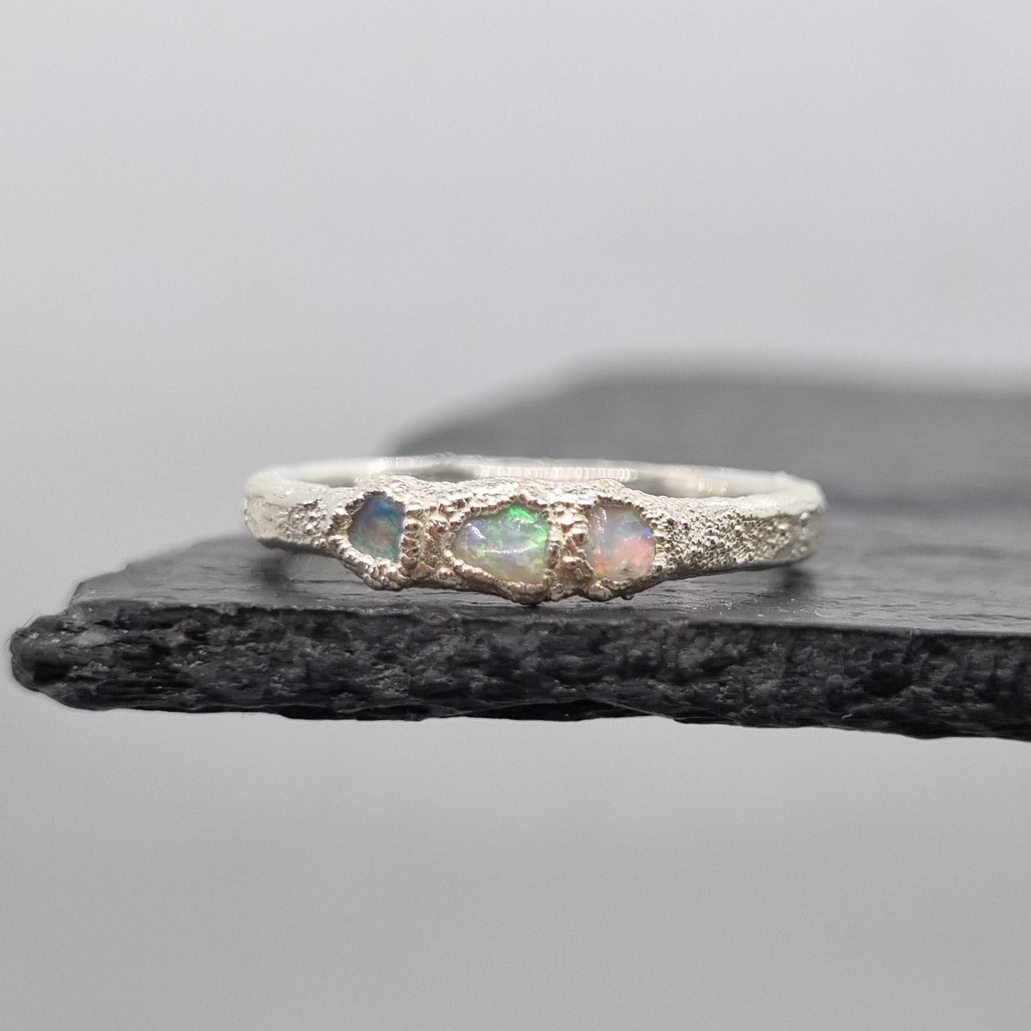 A silver ring featuring three small iridescent opal gemstones, displayed vertically against a dark textured slate on a light gray background.