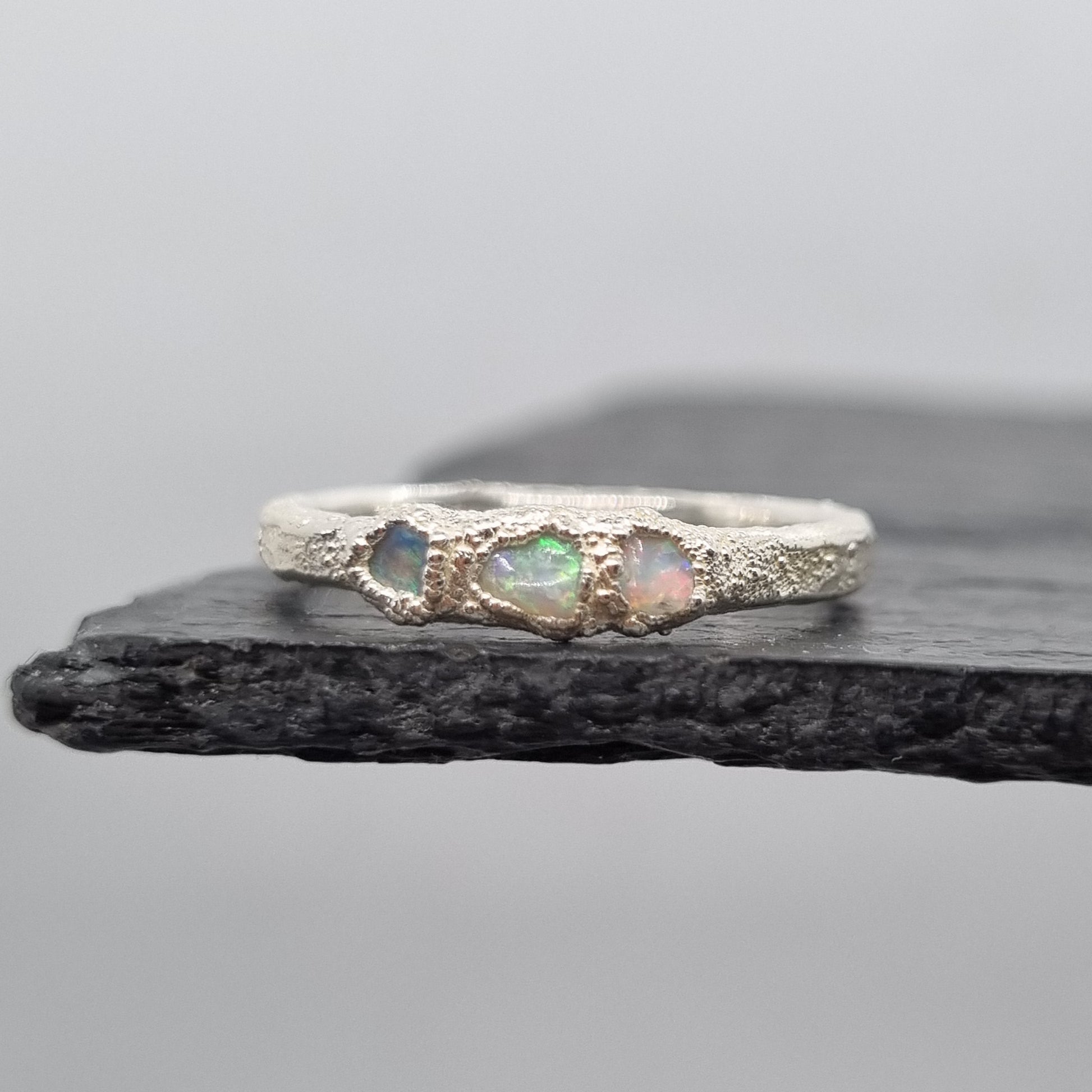 A silver ring featuring three small iridescent opal gemstones, displayed vertically against a dark textured slate on a light gray background.