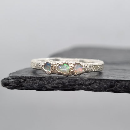 A silver ring featuring three small iridescent opal gemstones, displayed vertically against a dark textured slate on a light gray background.