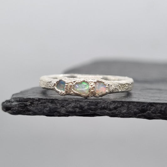 A silver ring featuring three small iridescent opal gemstones, displayed vertically against a dark textured slate on a light gray background.