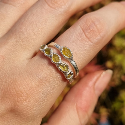 A close-up of a hand wearing two silver rings with raw yellow sapphires, set against a blurred outdoor background.
